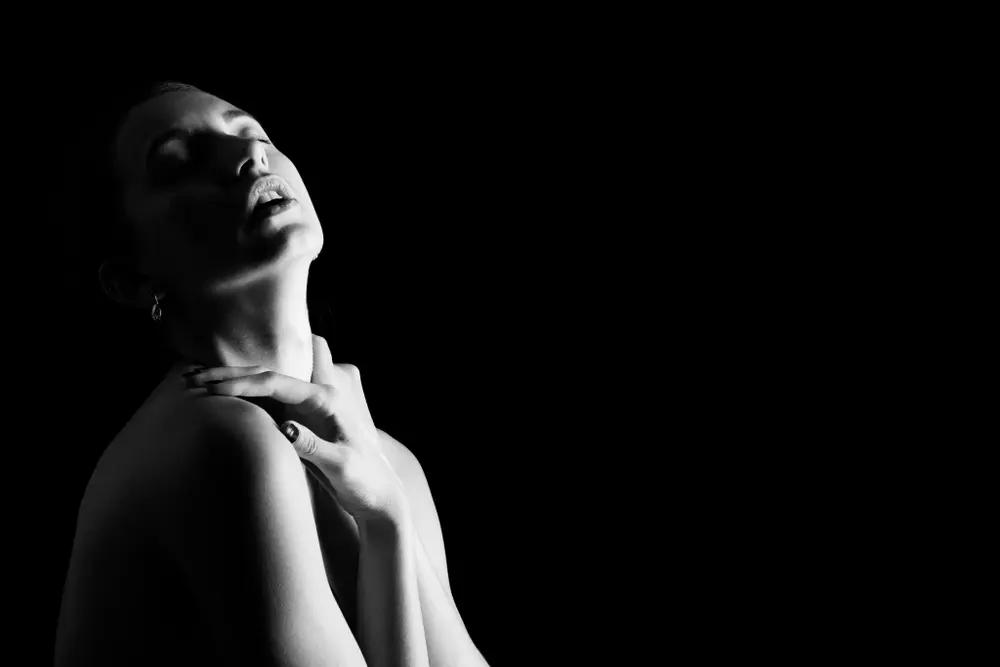 Monochrome fine-art portrait of young woman against black backdrop. Minimalist portrait photography exploring emotion.