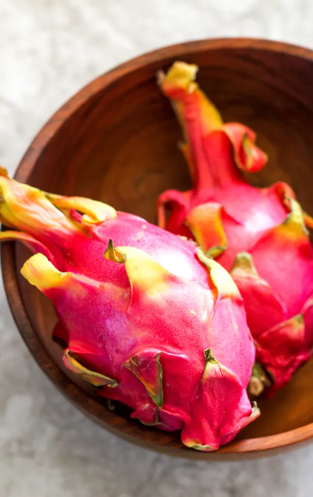 Two pink dragon fruits in a wooden bowl on a light stone background.