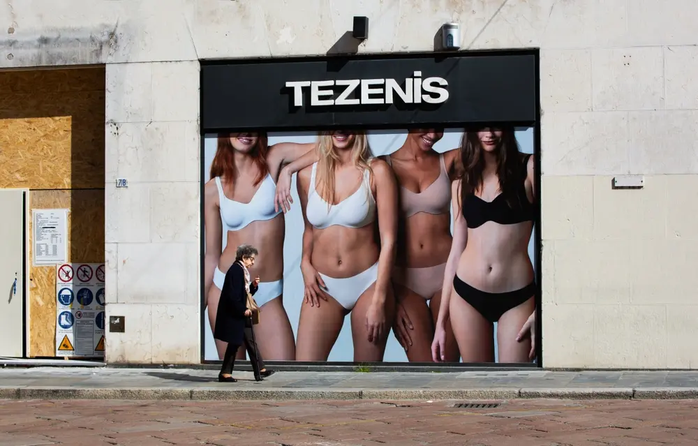 Candid street photograph, old woman walking past advertisement for women's underwear. Ironic juxtaposition in street photography.