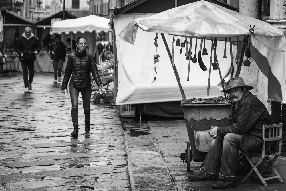 Rainy outdoor market scene. Old shopkeeper facing camera, seated.