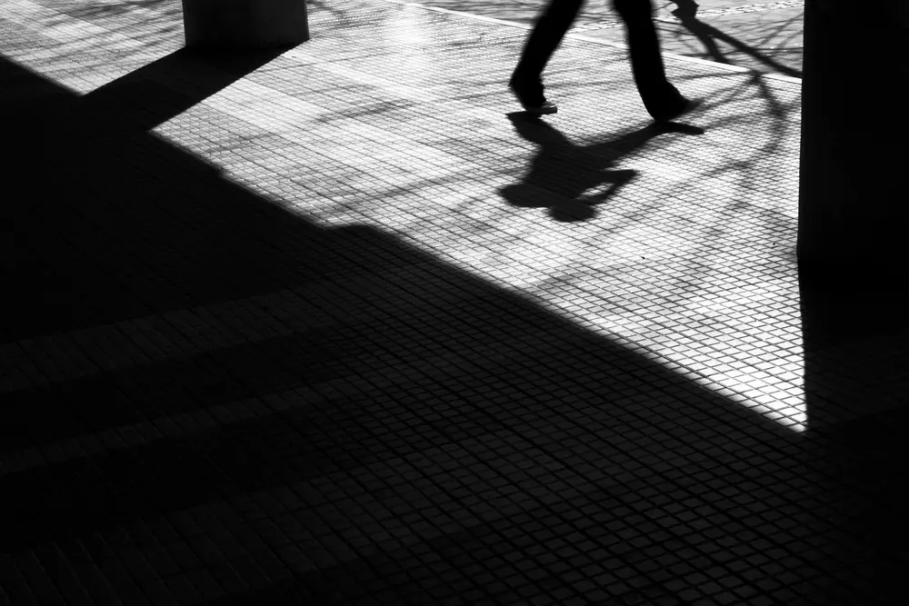 Low-perspective street photography of pedestrians walking against minimalist black-and-white backdrop. Harsh contrasts.