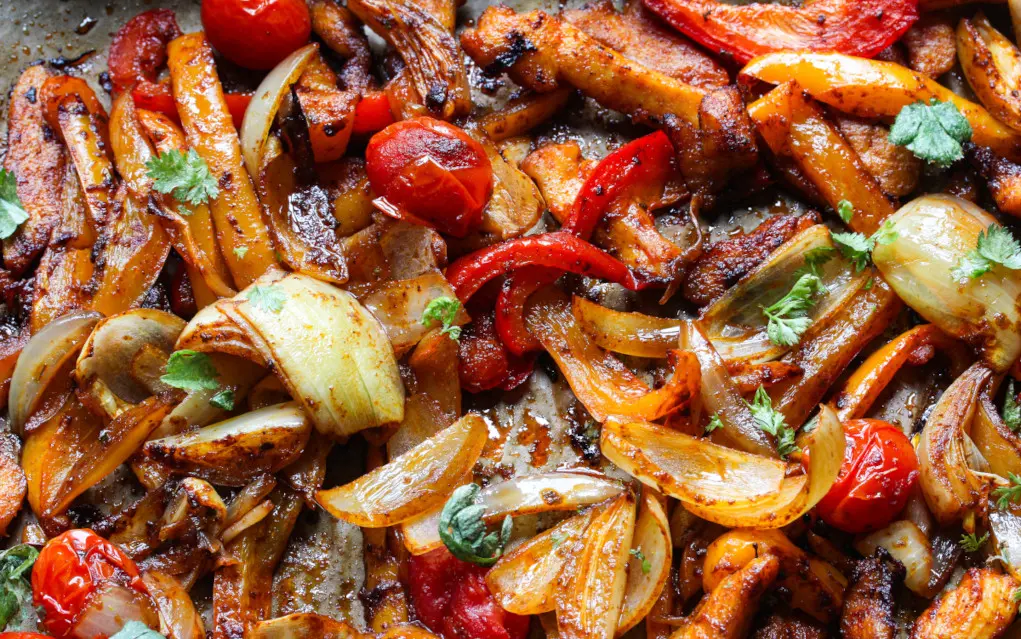 Colorful roasted peppers, onions, and tomatoes on a sheet pan glistening under bright lighting.