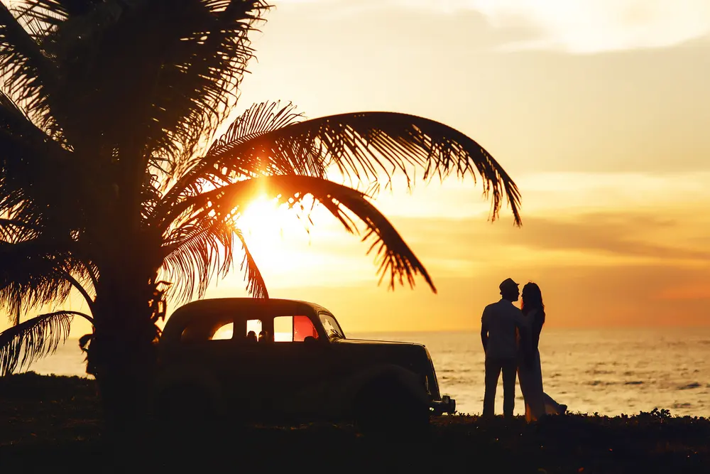 Romantic photograph of couple embracing at sunset. Vintage car, palm tree, and beachside scenery.