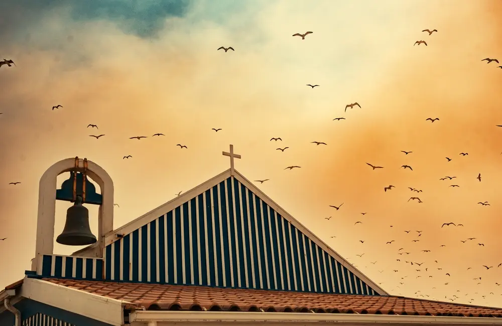 A flock of birds flying over the rooftop of a church. Emotional street photography shot from an upward angle.