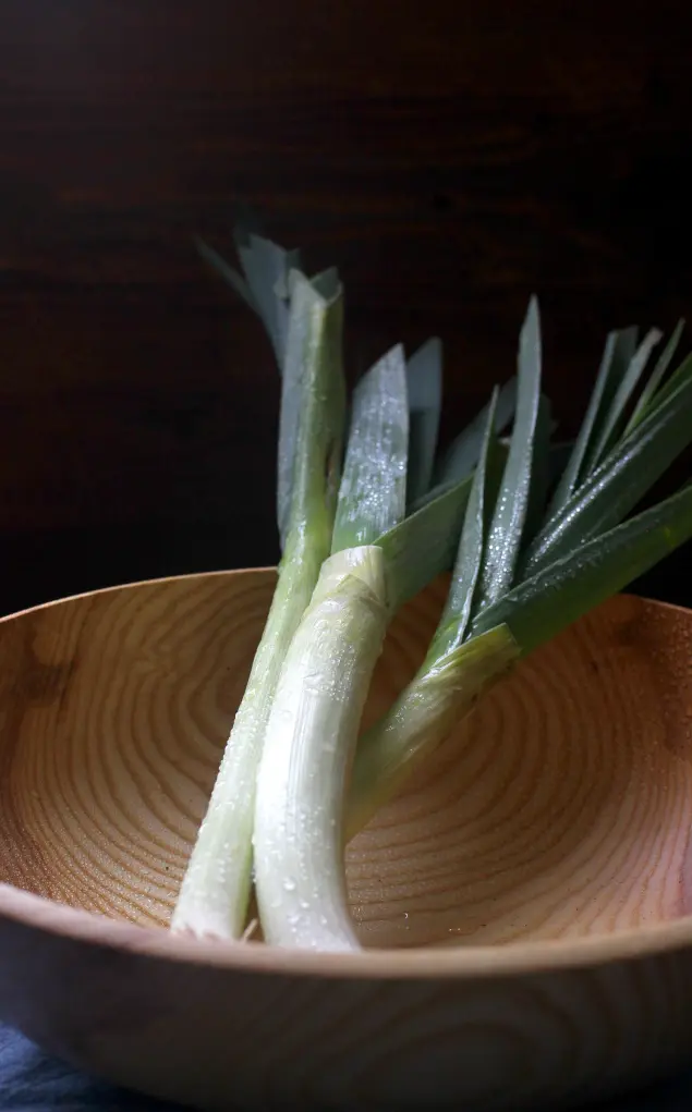  Fresh leeks with water droplets in a wooden bowl, lit by soft natural light.