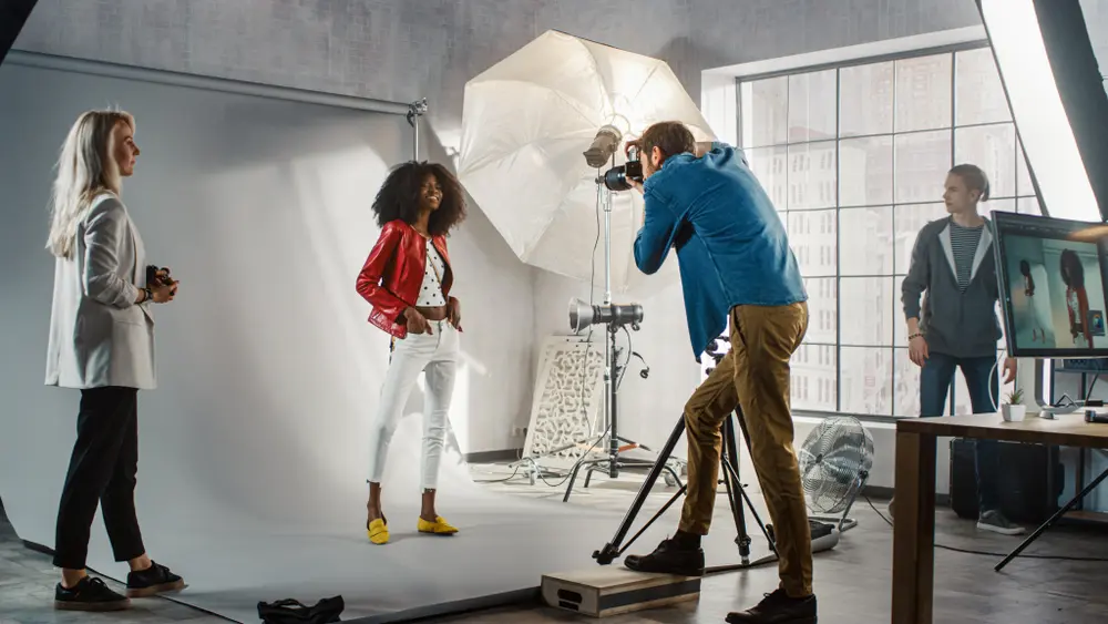 A studio fashion photographer composing a portrait of a young woman in a red coat against a white backdrop.