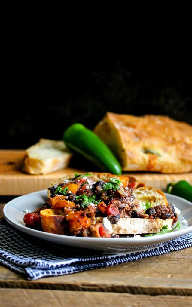 Plate of rustic vegetable bruschetta topped with herbs, photographed in warm golden-hour light with bread and jalapeños in the background.