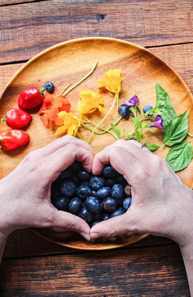 Hands forming a heart shape around a pile of fresh blueberries on a wooden plate with colorful edible flowers and tomatoes.