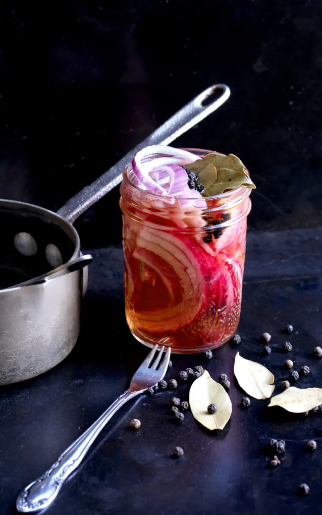 Jar of pickled onions with bay leaves and peppercorns, styled with a fork and saucepan in the background.
