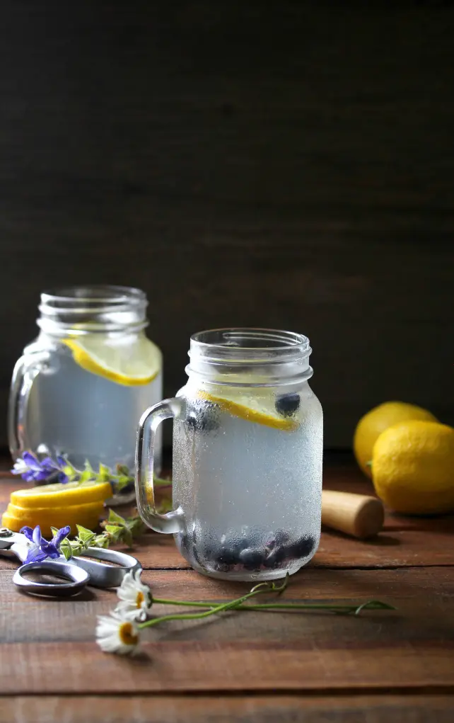 Lemon and blueberry-infused water in mason jars with flowers on a wooden table, photographed in soft natural light on a wooden table.