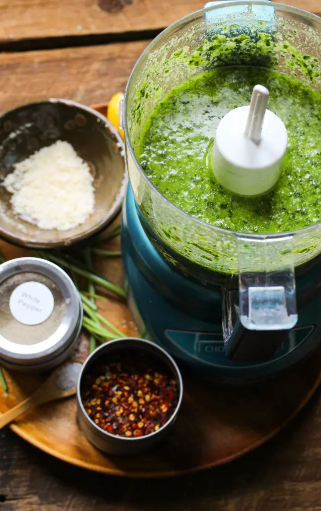 Fresh basil pesto in a food processor surrounded by Parmesan cheese, chili flakes, and spices, photographed using the rule of thirds.