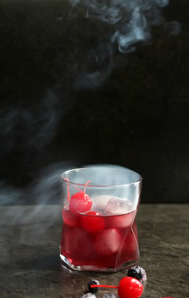 Smoked cocktail with cherries and ice in a glass, surrounded by drifting tendrils of smoke.