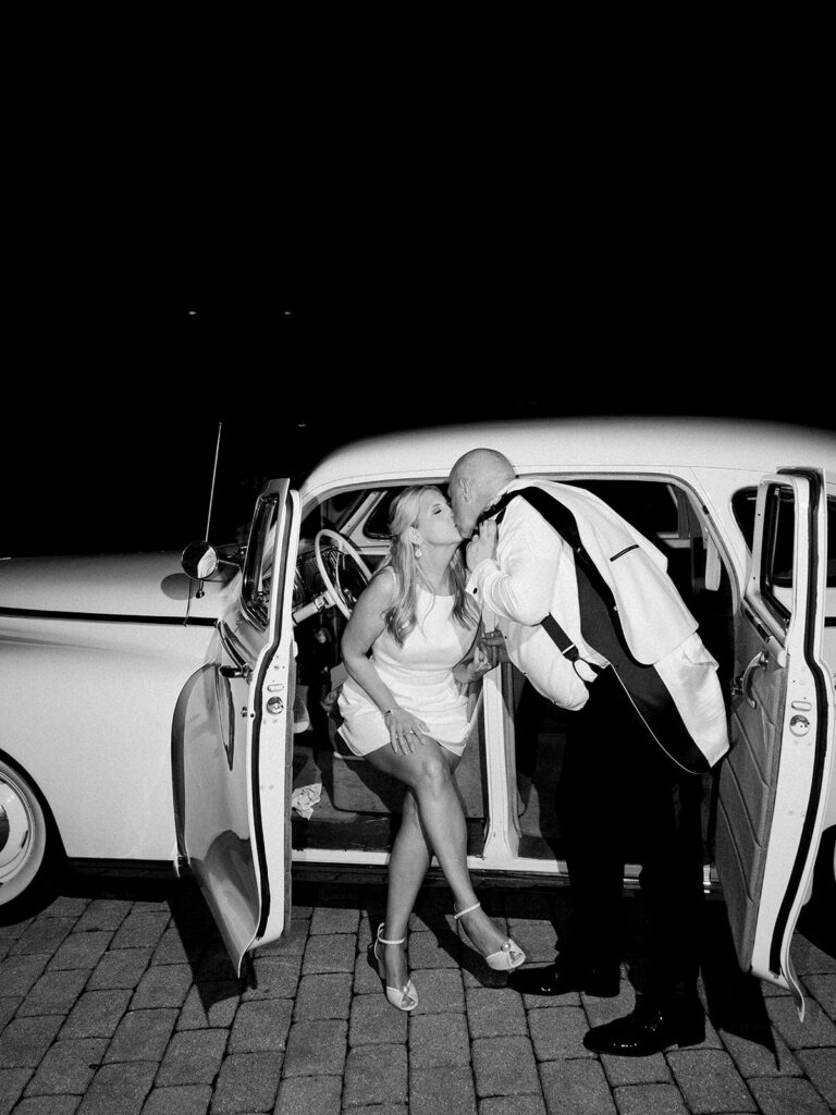 A black and white image of a couple kissing near a vintage car by Hannah Bjorndal.