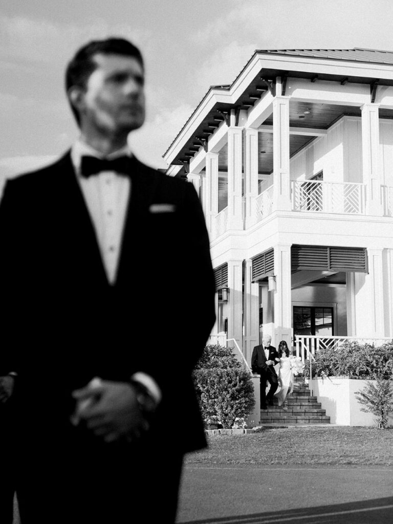 A black and white image of a groom out of focus in the foreground and a bride being walked towards him by her dad by Hannah Bjorndal