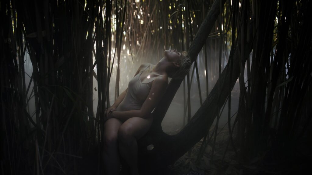 An image of a woman sitting and leaning against a tree in a densely forested area. 