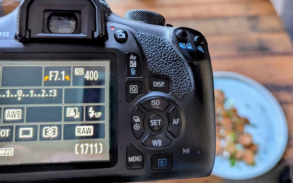 Close-up of the back of a DSLR camera showing its settings screen, with a blurred plate of food in the background.