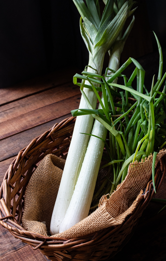 Fresh leeks with dewdrops arranged in a woven basket on a rustic wooden table, photographed in soft, moody light.