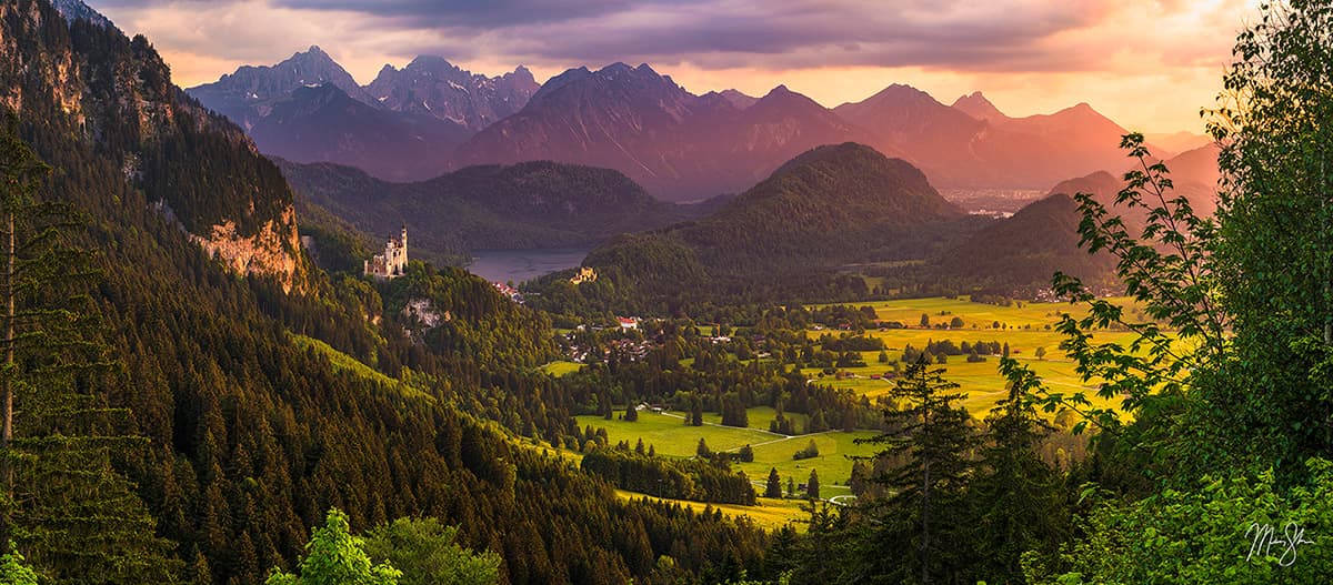 Sunlit valley view with Neuschwanstein Castle surrounded by forests and mountains in Bavaria.