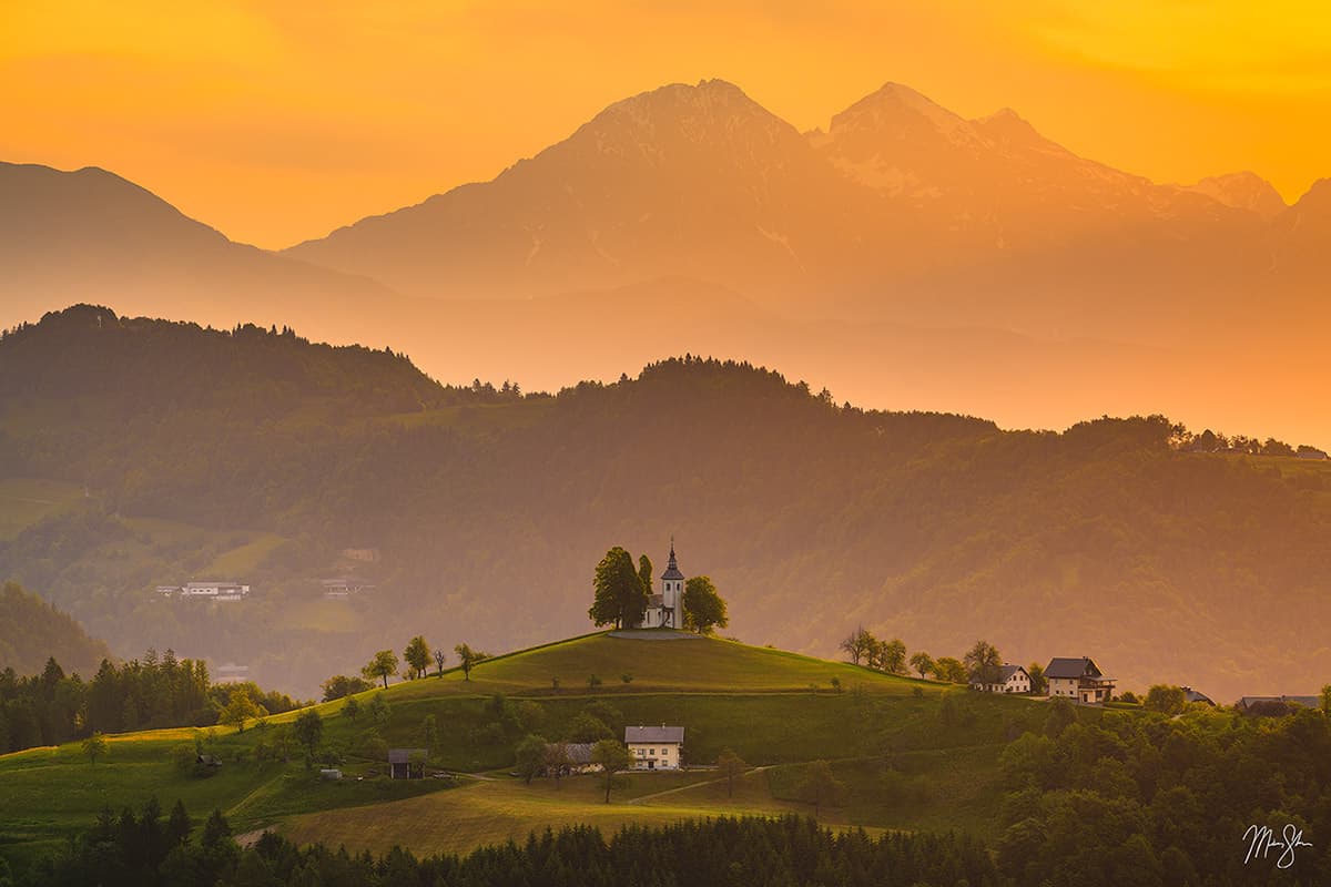 Hilltop church at sunrise with soft layers of mountains in rural Slovenia.