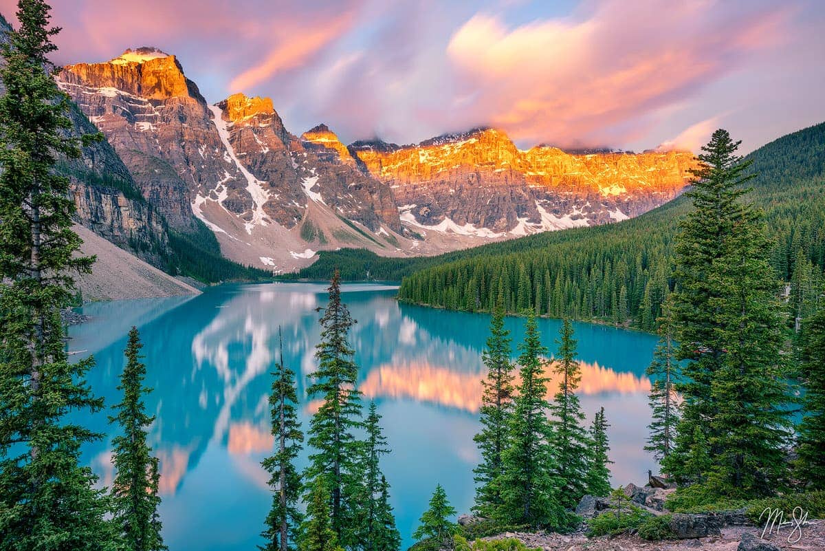 Sunrise light reflecting on Moraine Lake with pine trees and rugged peaks in Banff National Park.