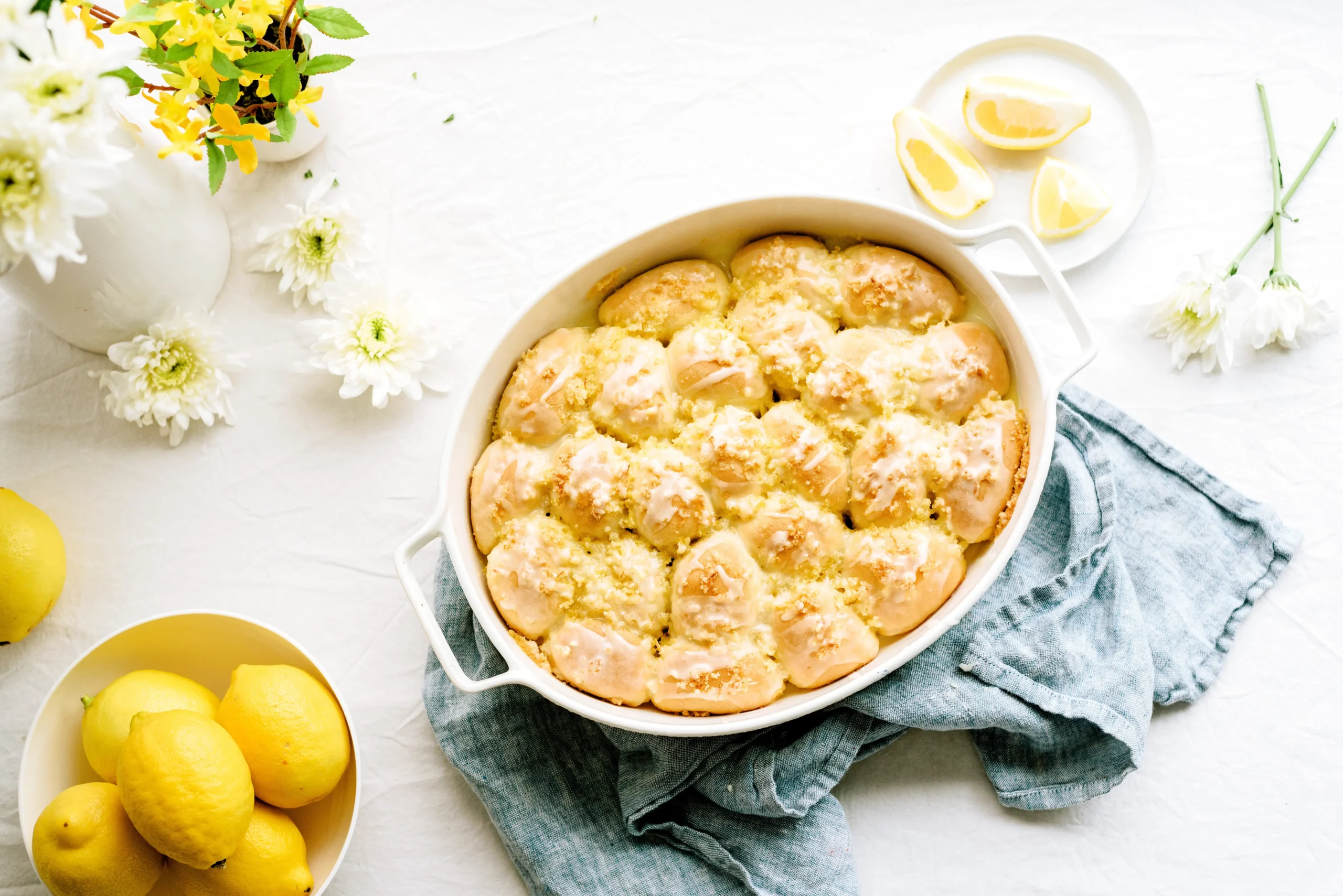 Overhead photograph of a lemon dessert in a baking dish styled with lemons, linens, flowers, and serving plates to demonstrate prop selection and scene design.