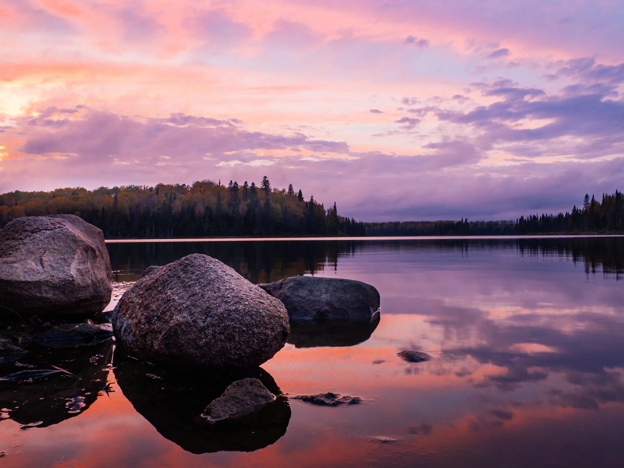 Large rocks in the foreground of a calm lake at sunset, creating depth and guiding the viewer's eye toward the distant treeline and colorful sky.