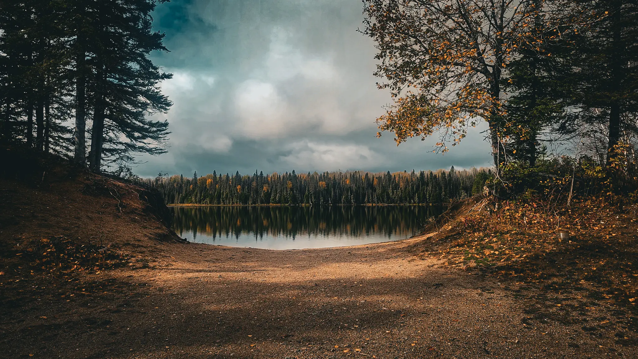Trees on both sides frame a calm reflective lake, creating a symmetrical landscape scene viewed from the sandy shoreline.