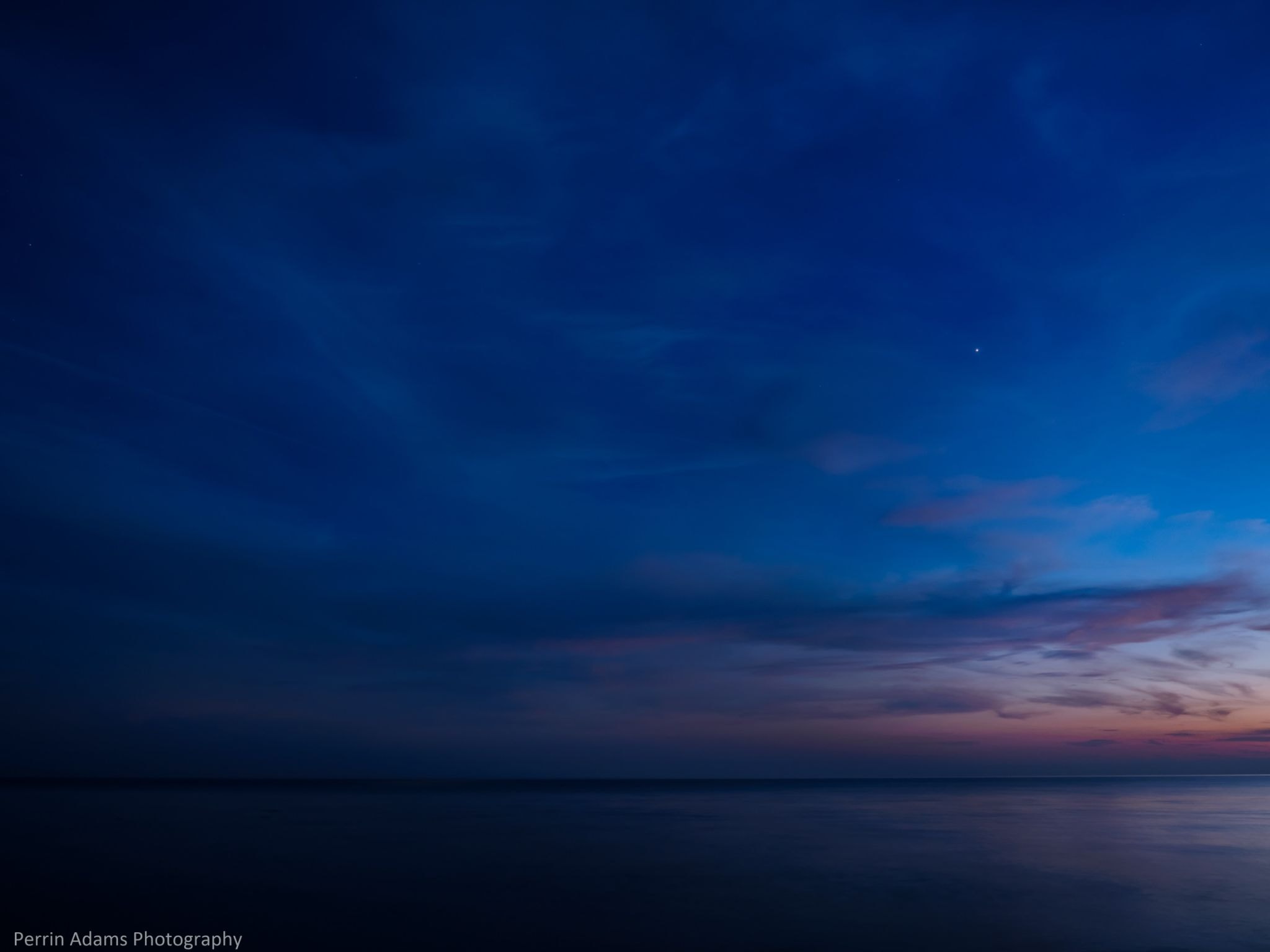 A calm shoreline at blue hour with deep blue sky, faint sunset colors on the horizon, and soft, low-contrast lighting across the water.