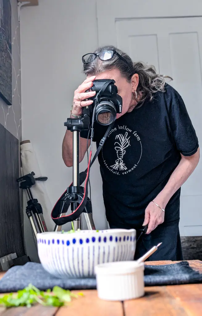 Woman adjusting a DSLR camera on a tripod while shooting a food scene on a table.