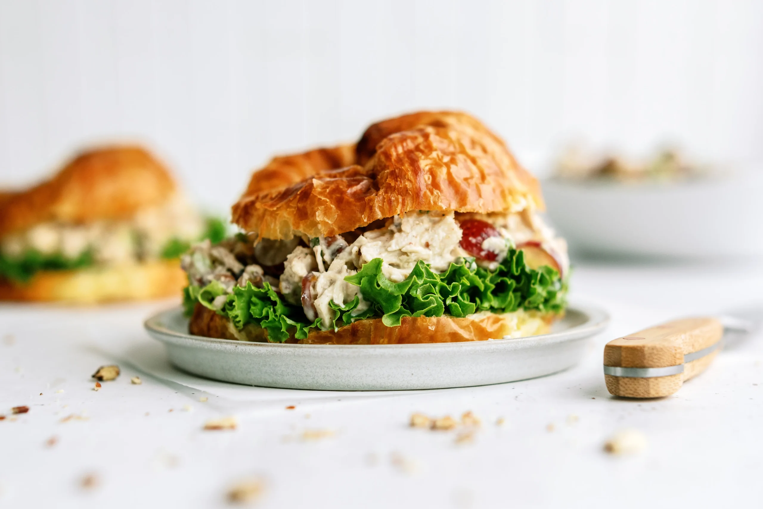 Layered photograph of a croissant sandwich with textured fillings, crumbs, and background elements creating depth and story.
