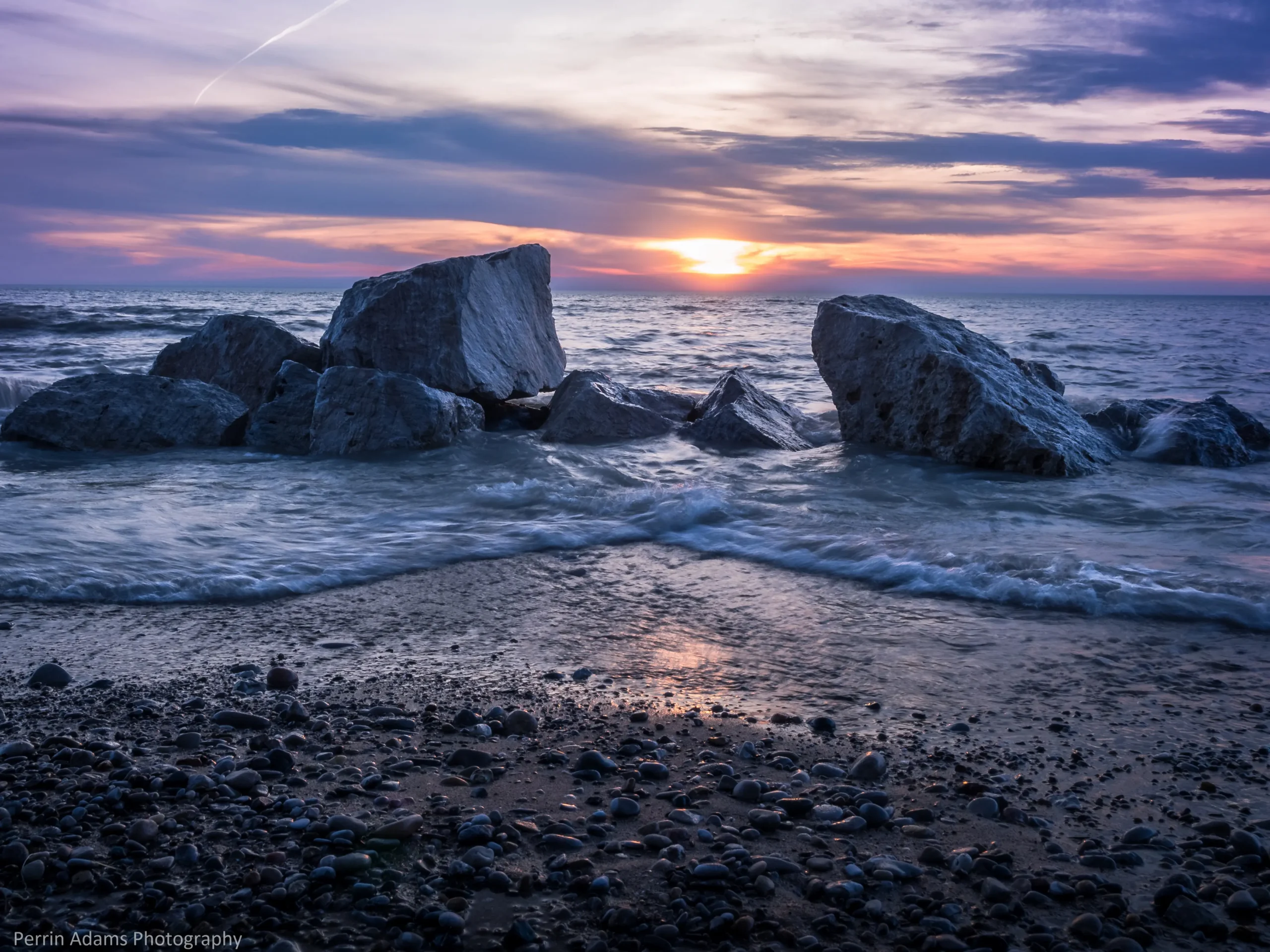Sunset over a rocky shoreline, with large boulders in the surf and a pebble-covered beach in the foreground beneath streaked clouds.