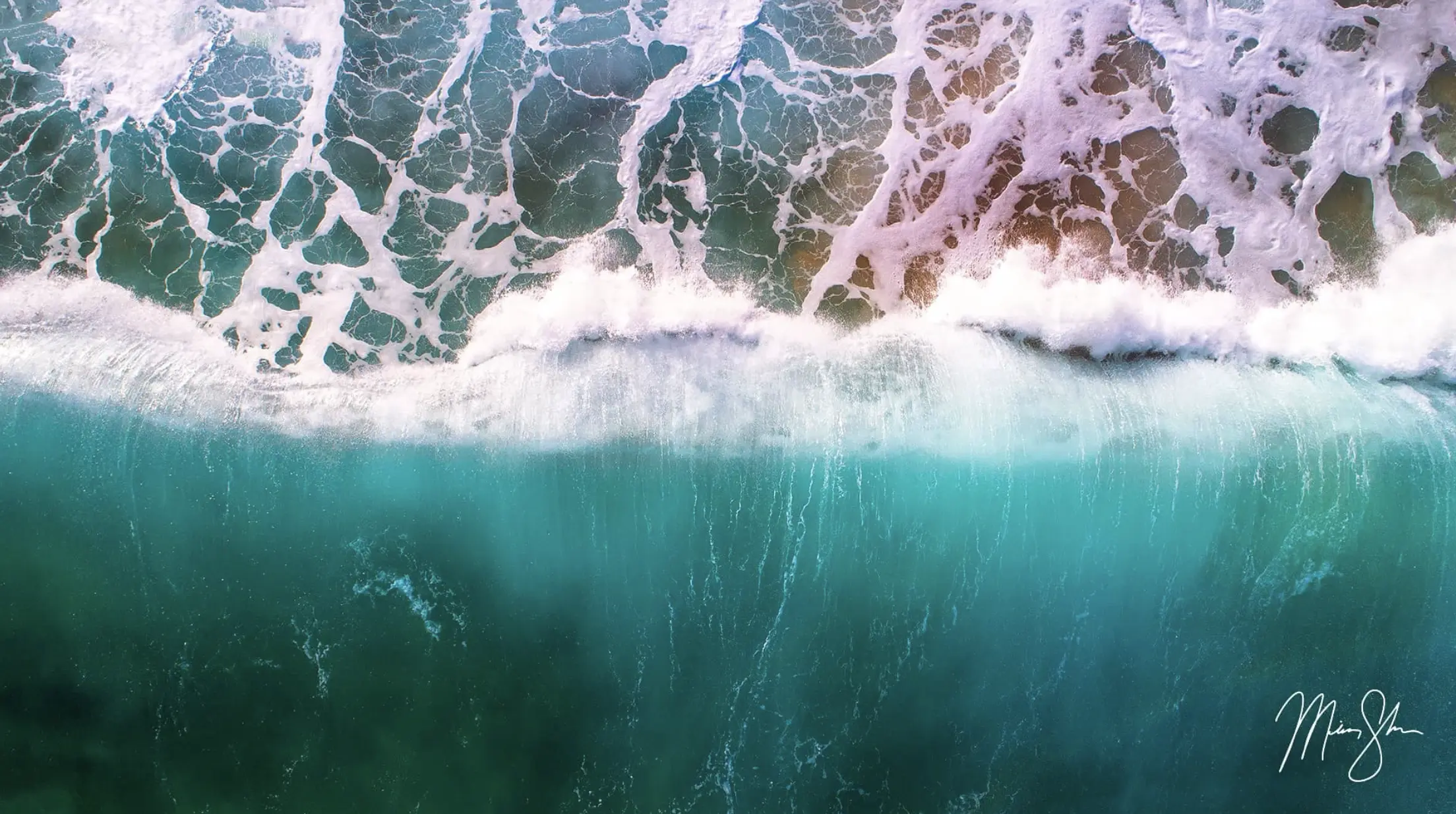 Aerial view of a turquoise ocean wave breaking with white sea foam patterns along the shoreline.