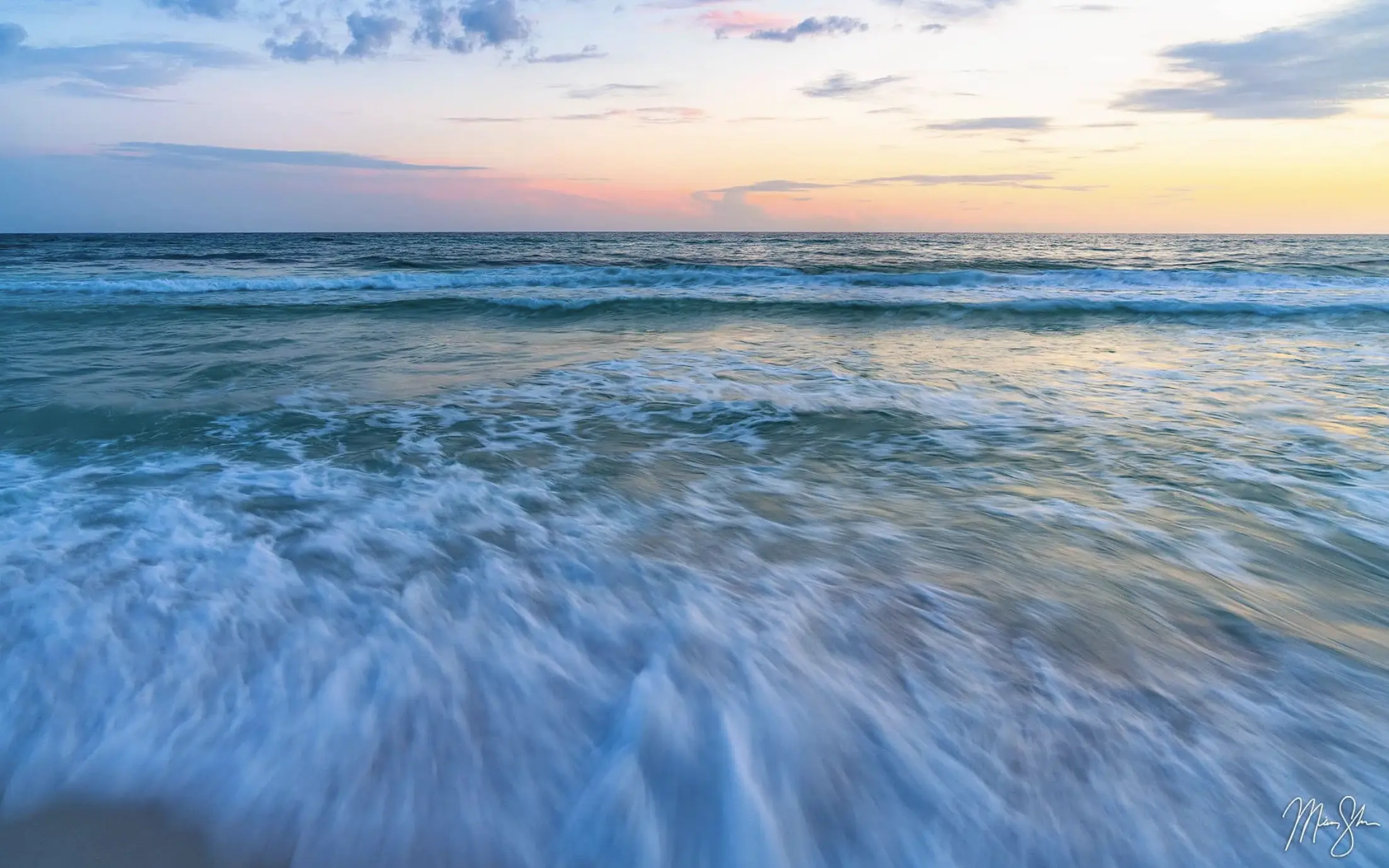 Soft waves washing toward the shore at sunset with a pastel sky and calm horizon.