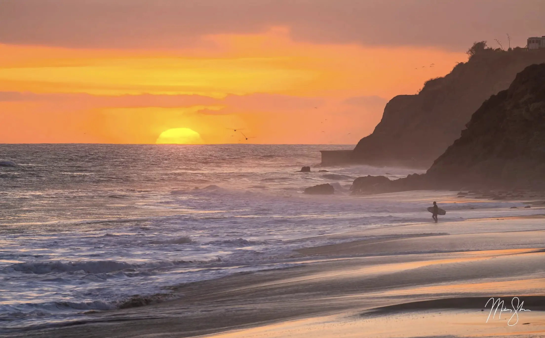 Surfer walking along a beach at sunset with waves rolling in and dark cliffs silhouetted against an orange sky.