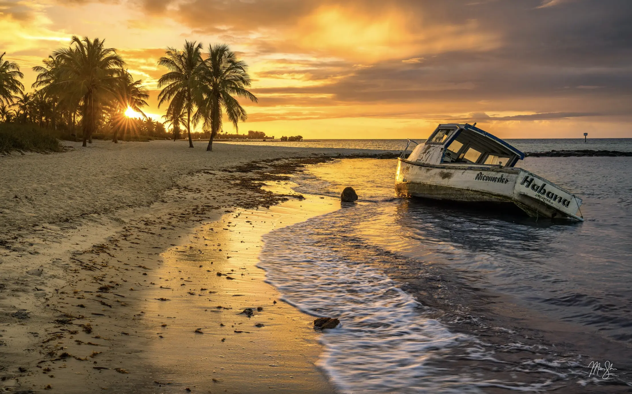 Abandoned boat on a sandy tropical beach at sunset with palm trees and golden reflections on the water.
