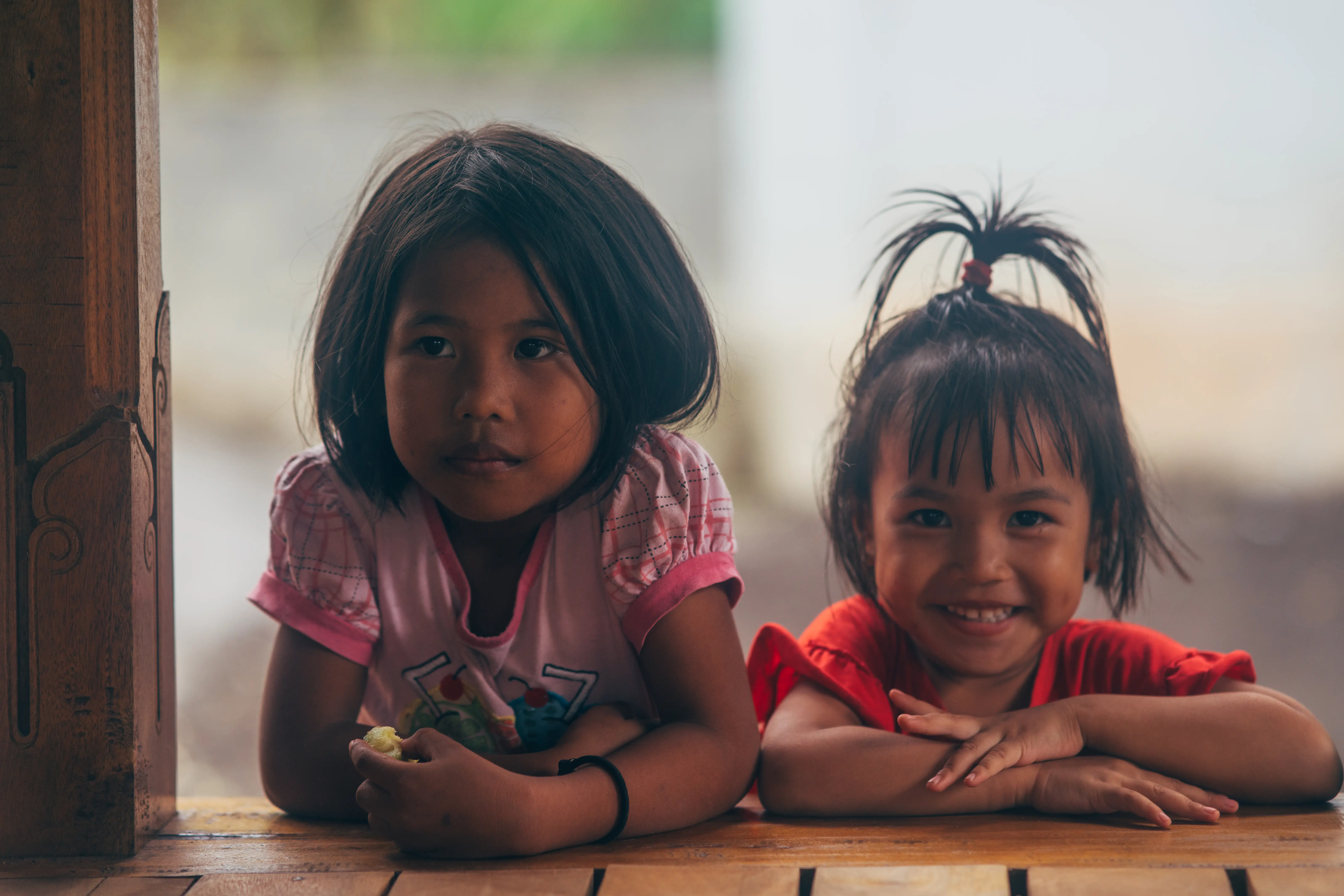 Two young girls leaning on a wooden window frame at a cassava farm in Indonesia, one smiling at the camera while the other looks thoughtfully to the side.