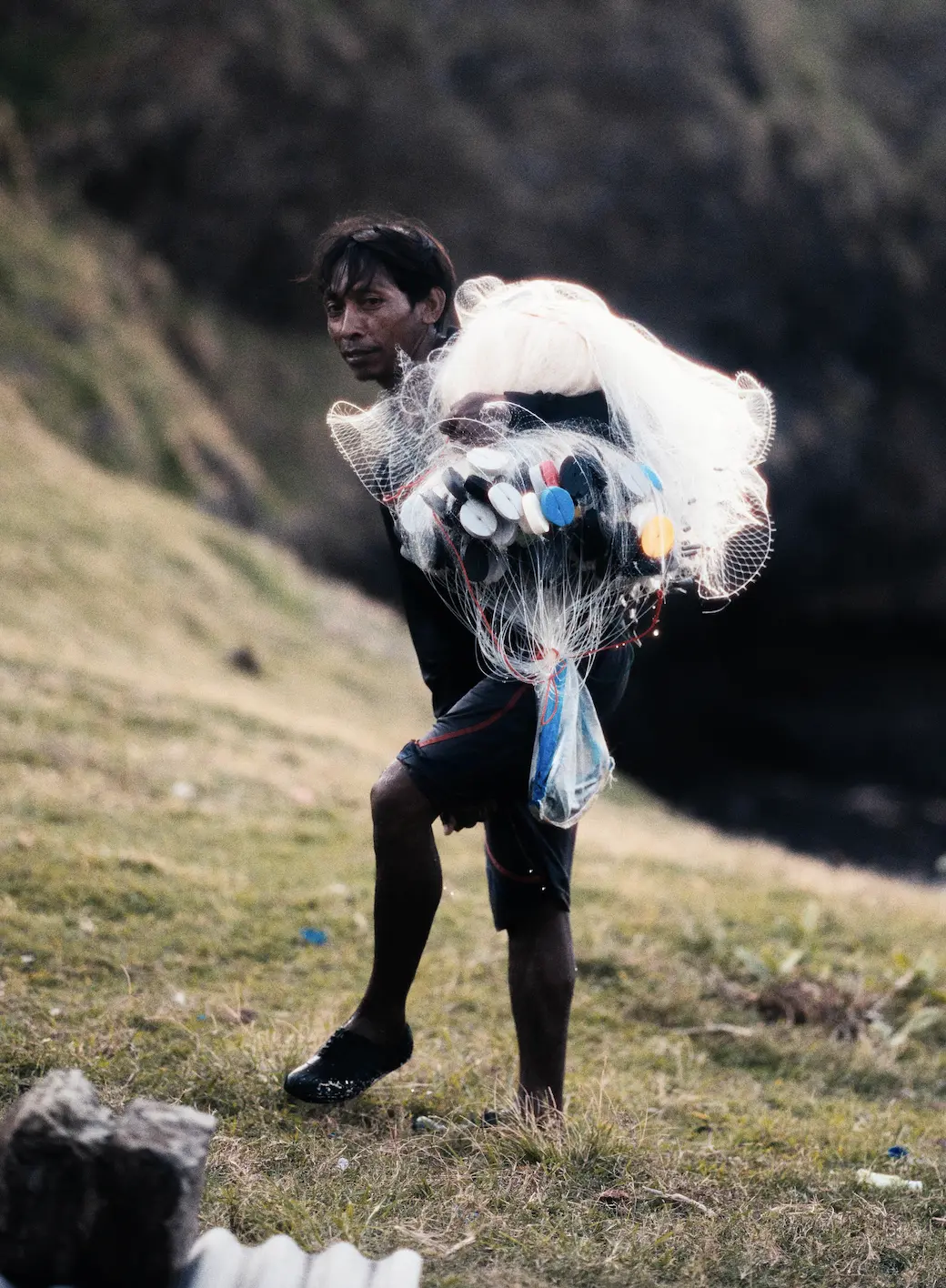 Fisherman walking on a grassy hillside carrying a bundle of fishing nets in Indonesia.