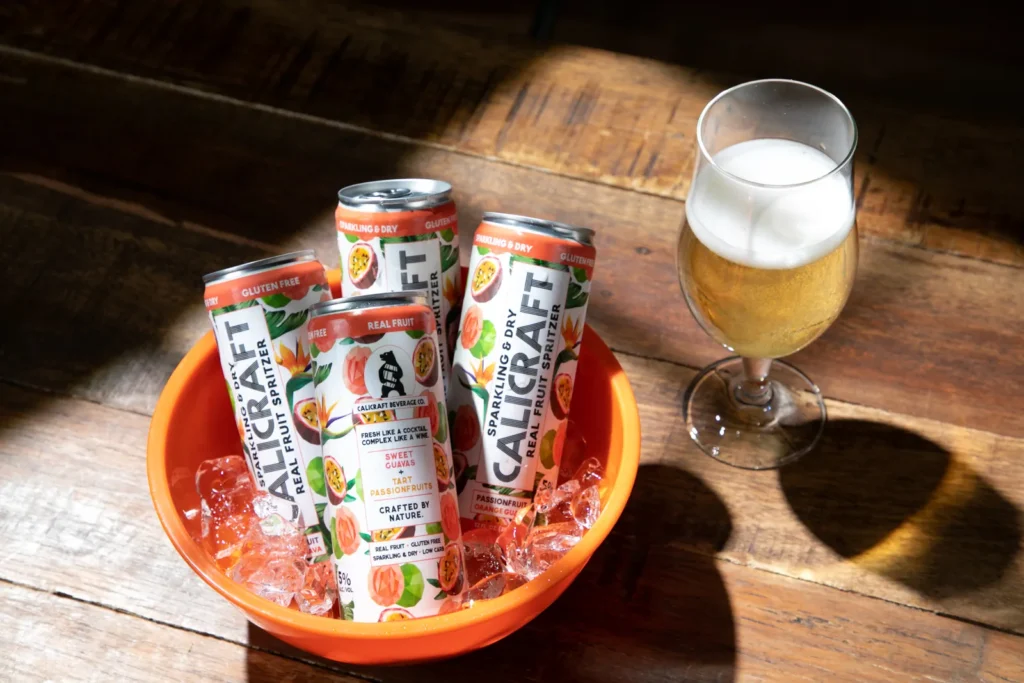 A photography of some cans of fruit spritzer sitting in a bow of ice with a glass beside it filled with some of the drink by Shelly Waldman.