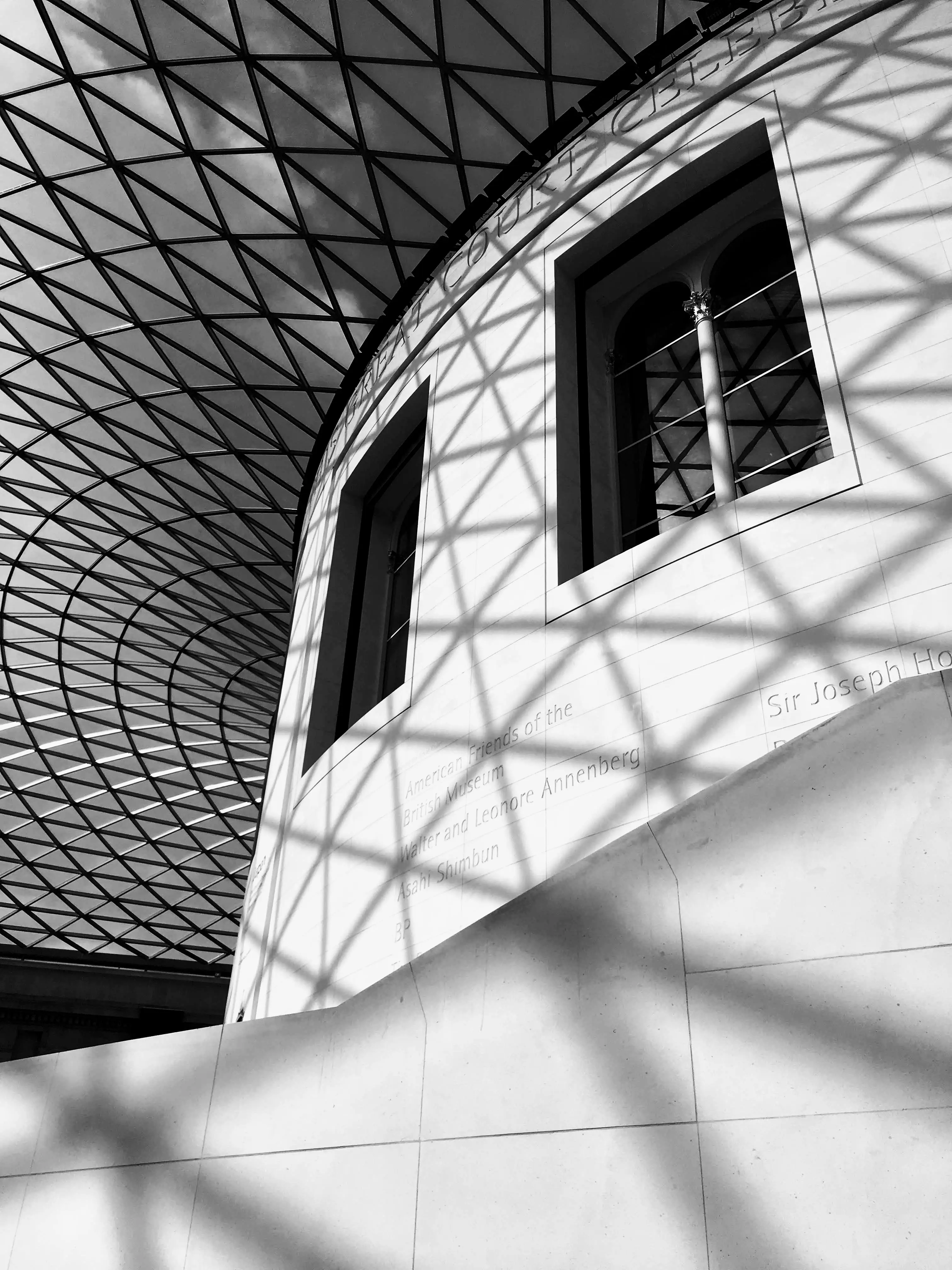 The Great Court of the British Museum, London with steel roof structure casting geometric shadows across the curved Reading Room wall.