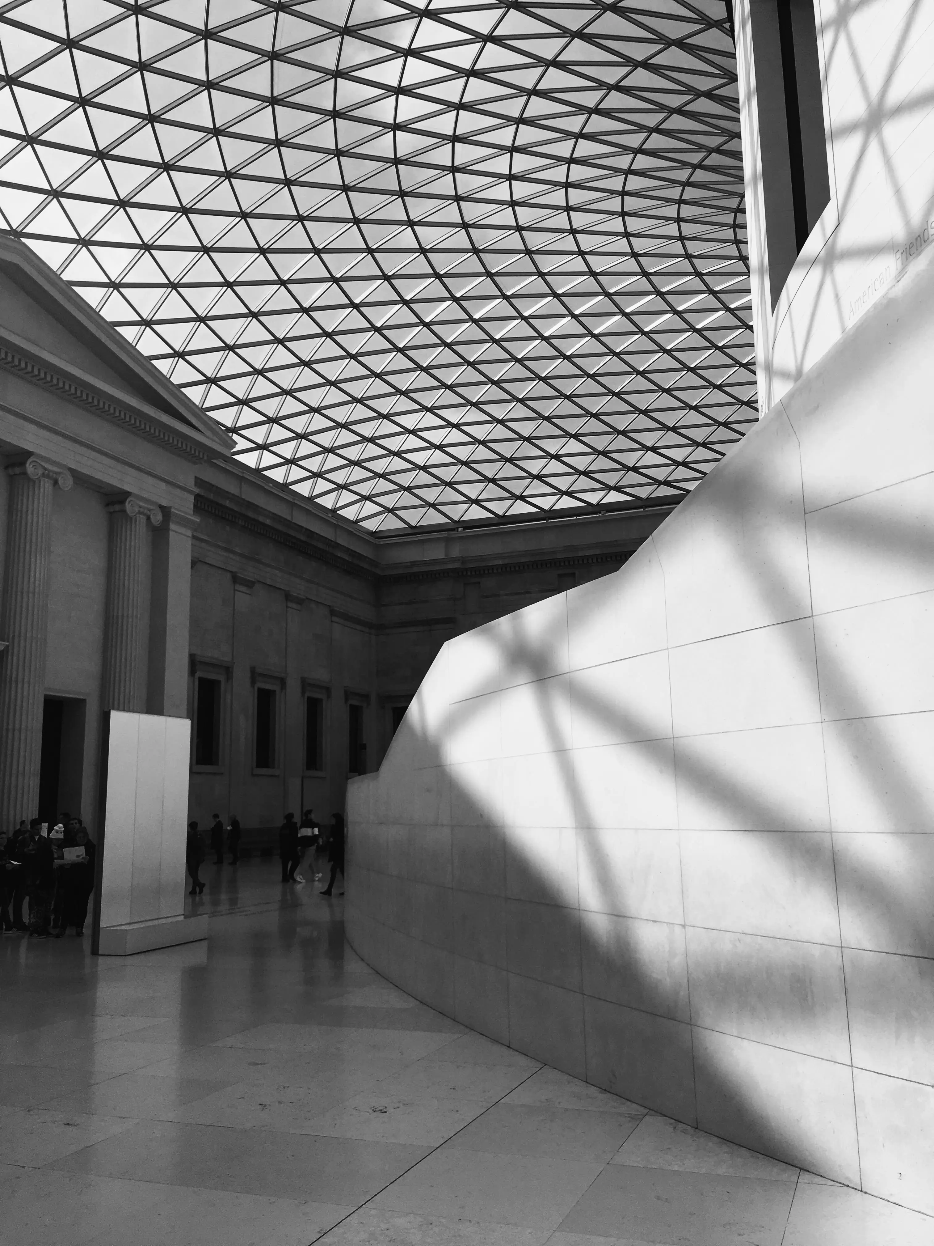 Harsh midday light casting geometric shadows from the steel roof grid across the curved wall of the British Museum Great Court, London.