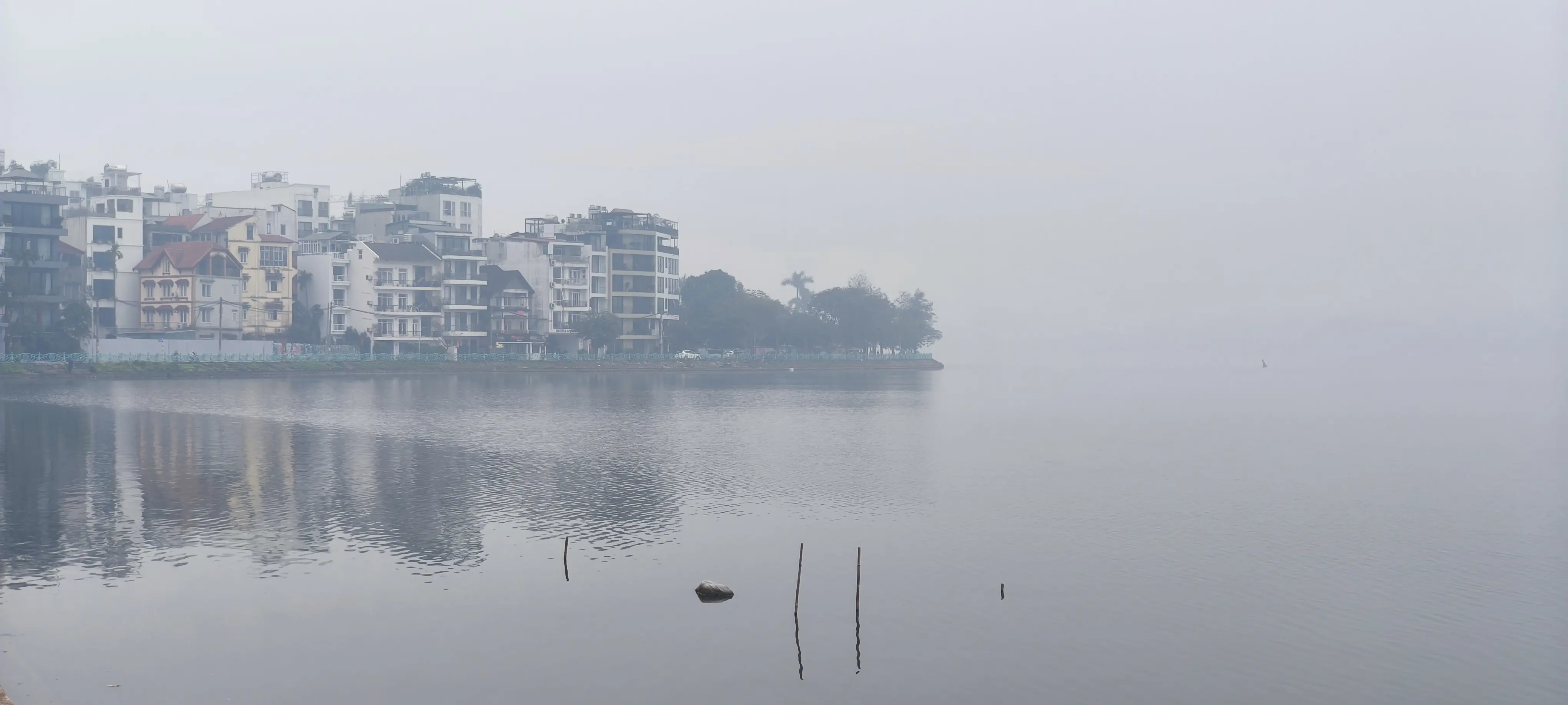 Misty morning on a Hanoi lake, buildings reflected in still water, illustrating the geometry of silence in urban architectural photography.
