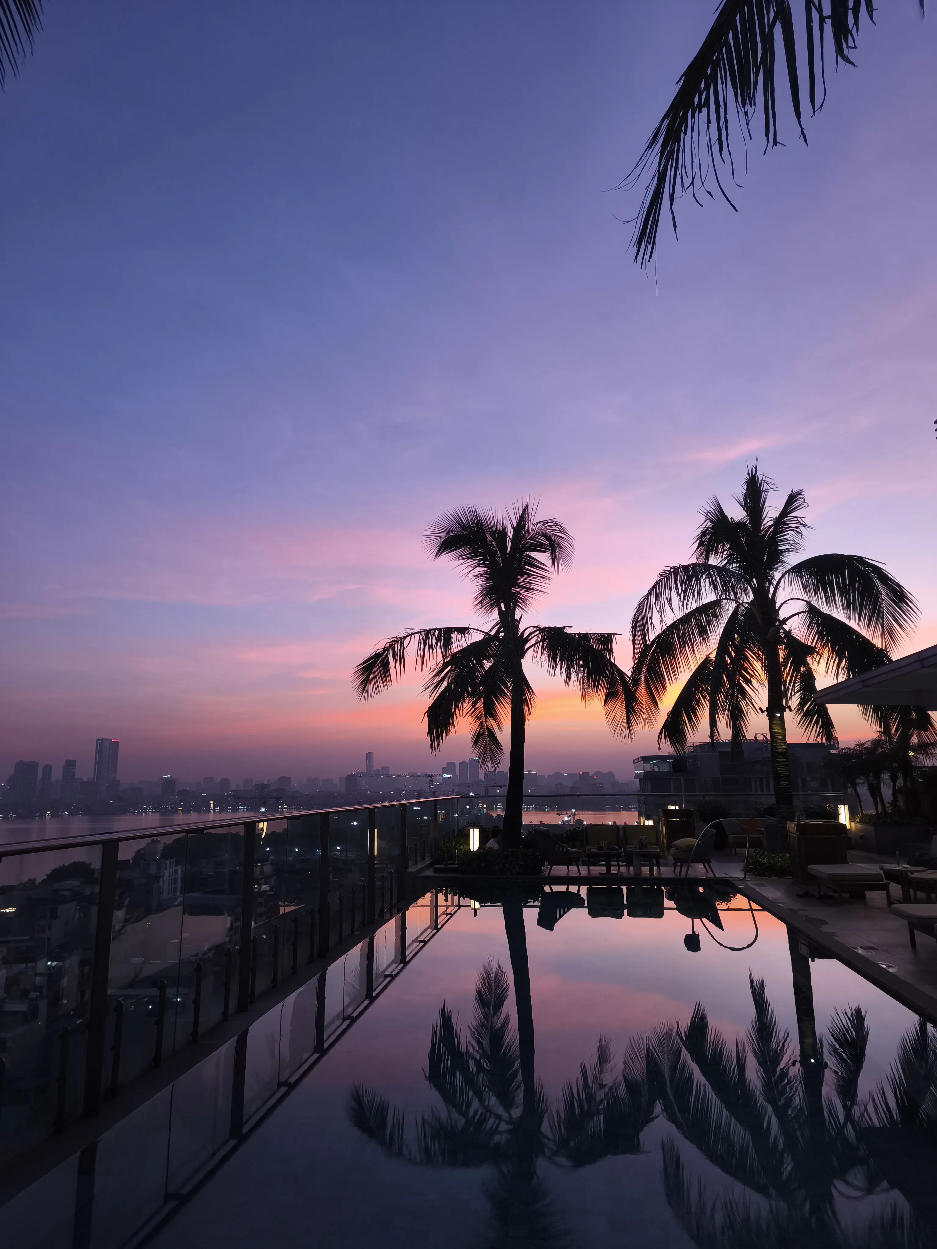 Blue hour view from a Hanoi rooftop infinity pool, palm tree silhouettes reflected in still water against a purple and orange dusk sky over the city skyline.