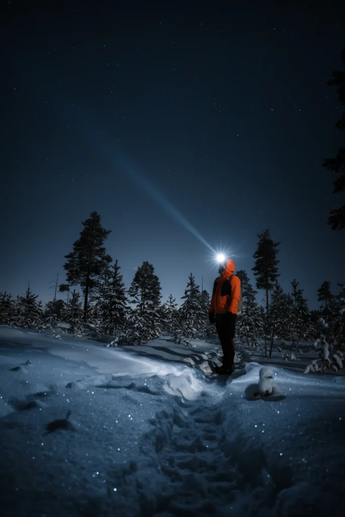 An image of a person standing in a winter forest with their headlight pointed up by Matti Seppänen
