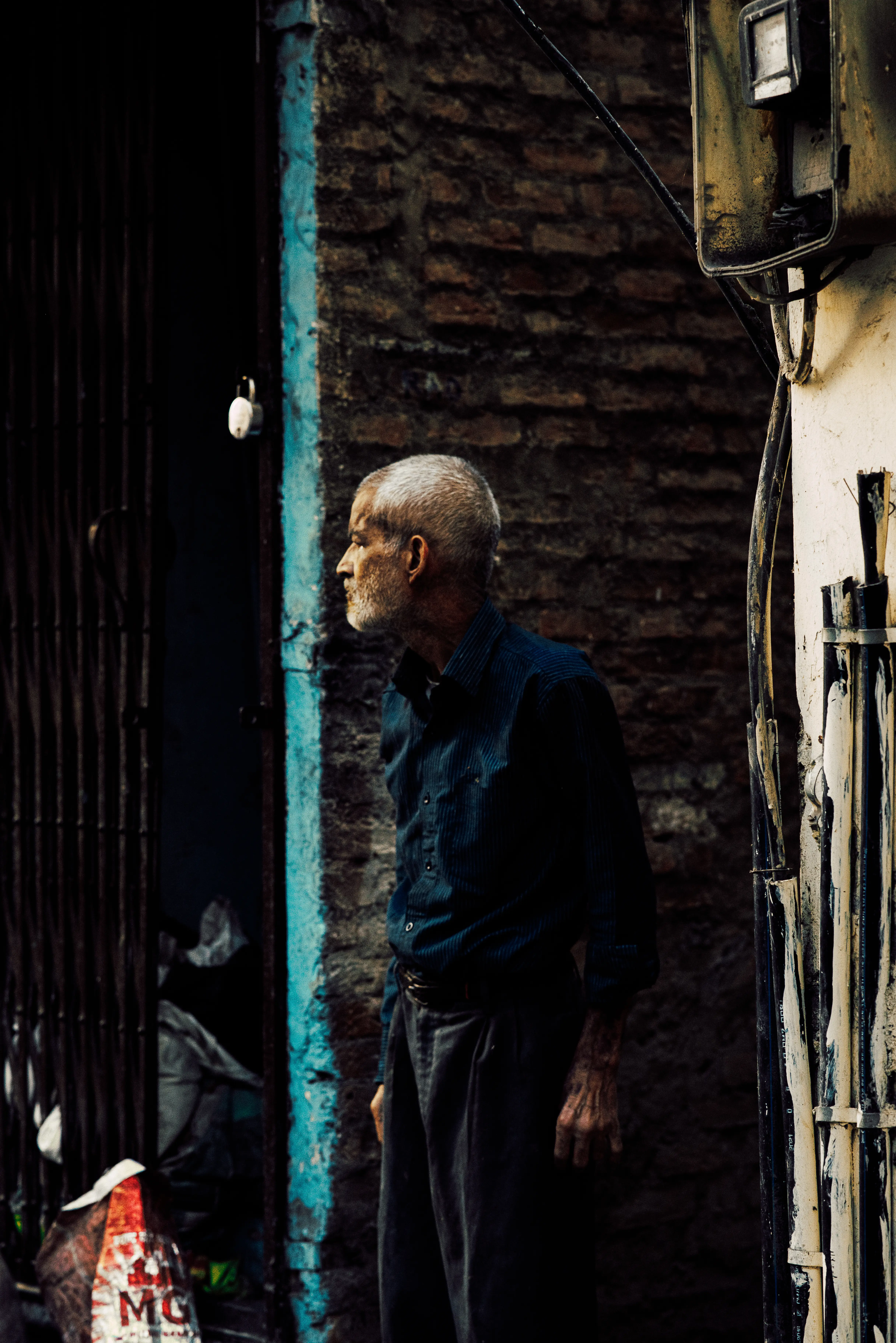 Grey-haired man in profile standing in a narrow alley, framed by vertical lines and lit by soft directional light off the walls
