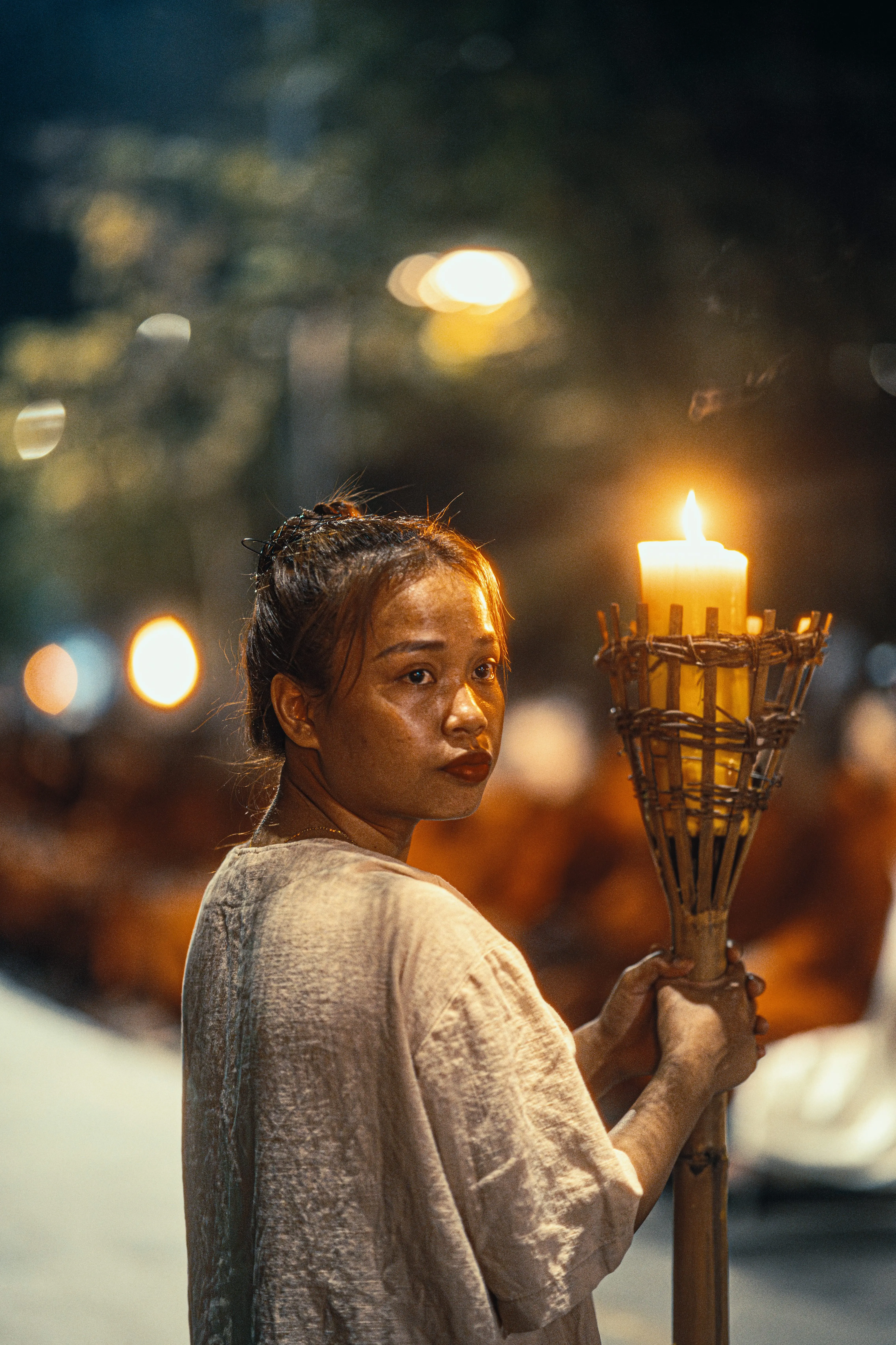 Young woman lit only by the candle torch she carries at a night ceremony, robed figures blurred behind her