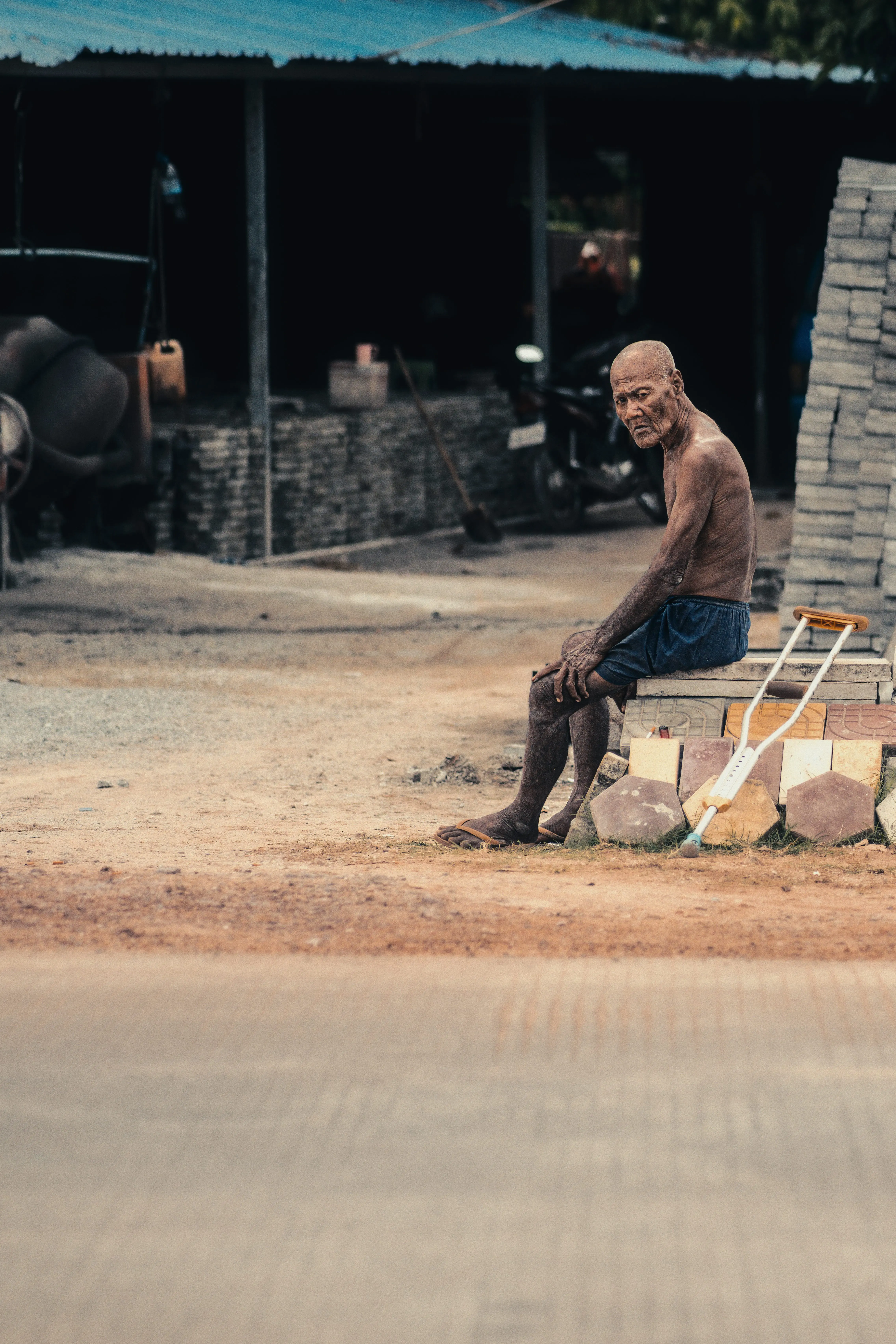 Shirtless elderly labourer sitting beside a crutch at a Cambodian construction site in harsh overhead afternoon light