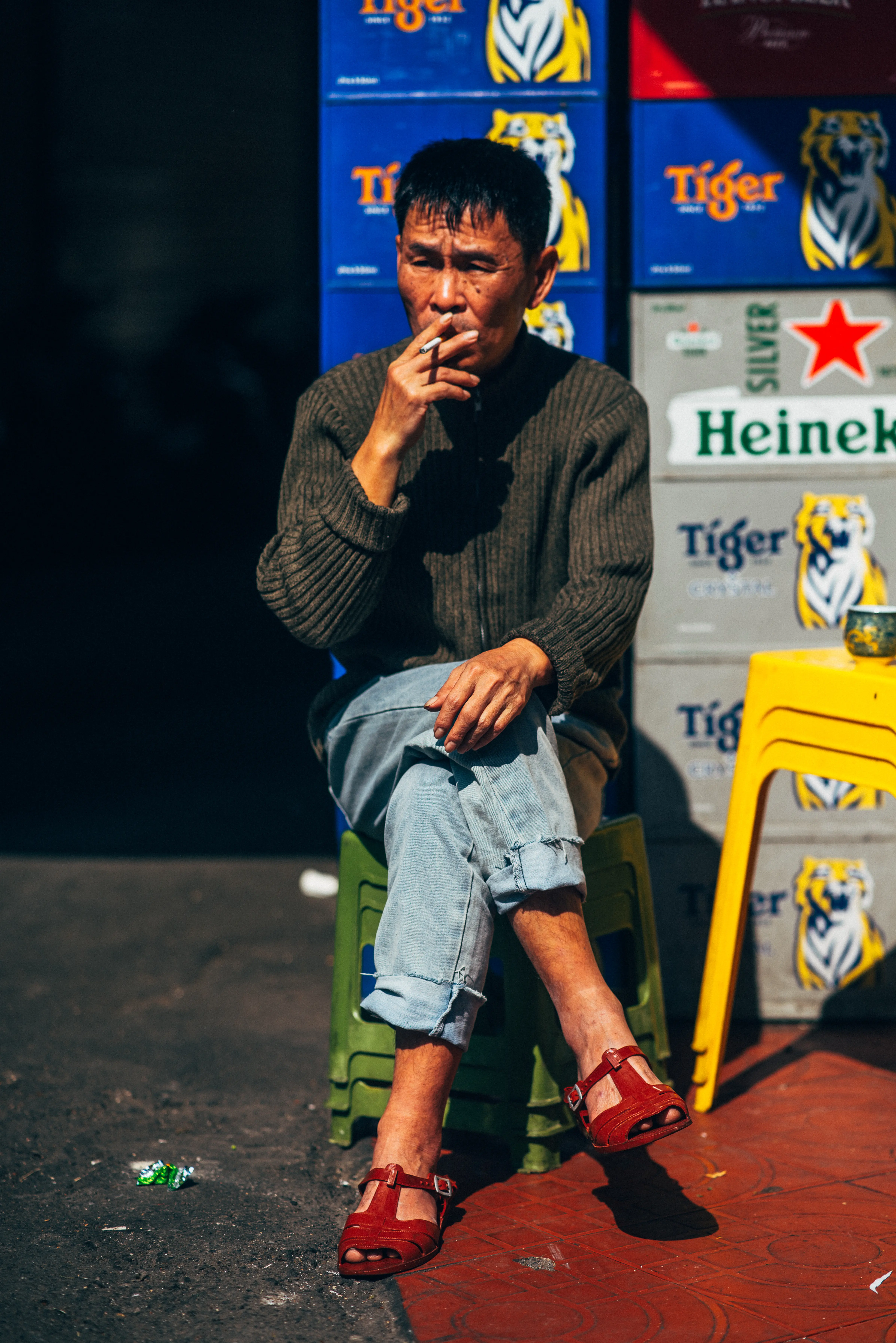 Man smoking on a plastic stool in harsh midday sun against stacked beer crates, half his face in shadow