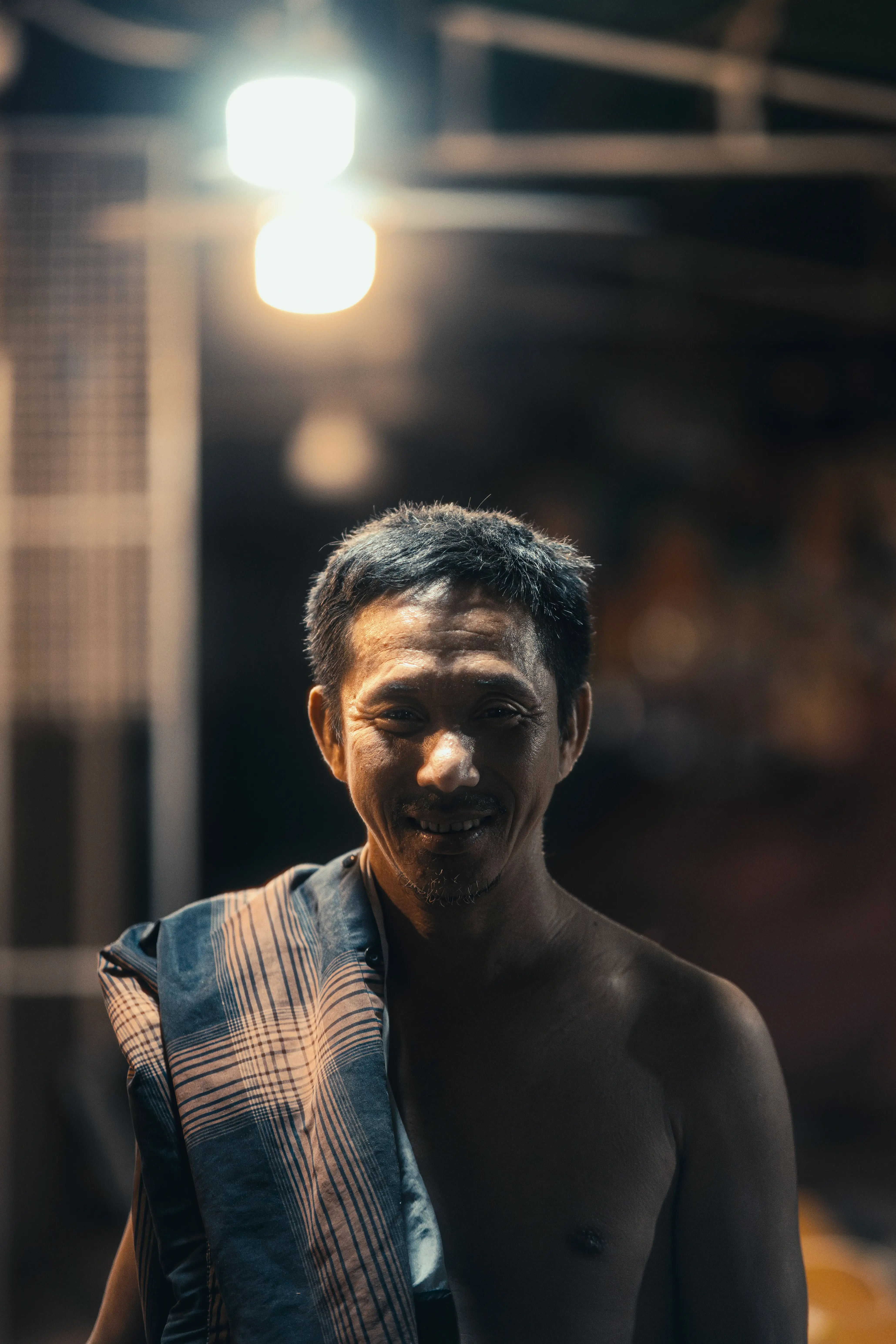 Man smiling beneath a single blown-out LED bulb at a Cambodian night market, face lit and background dark