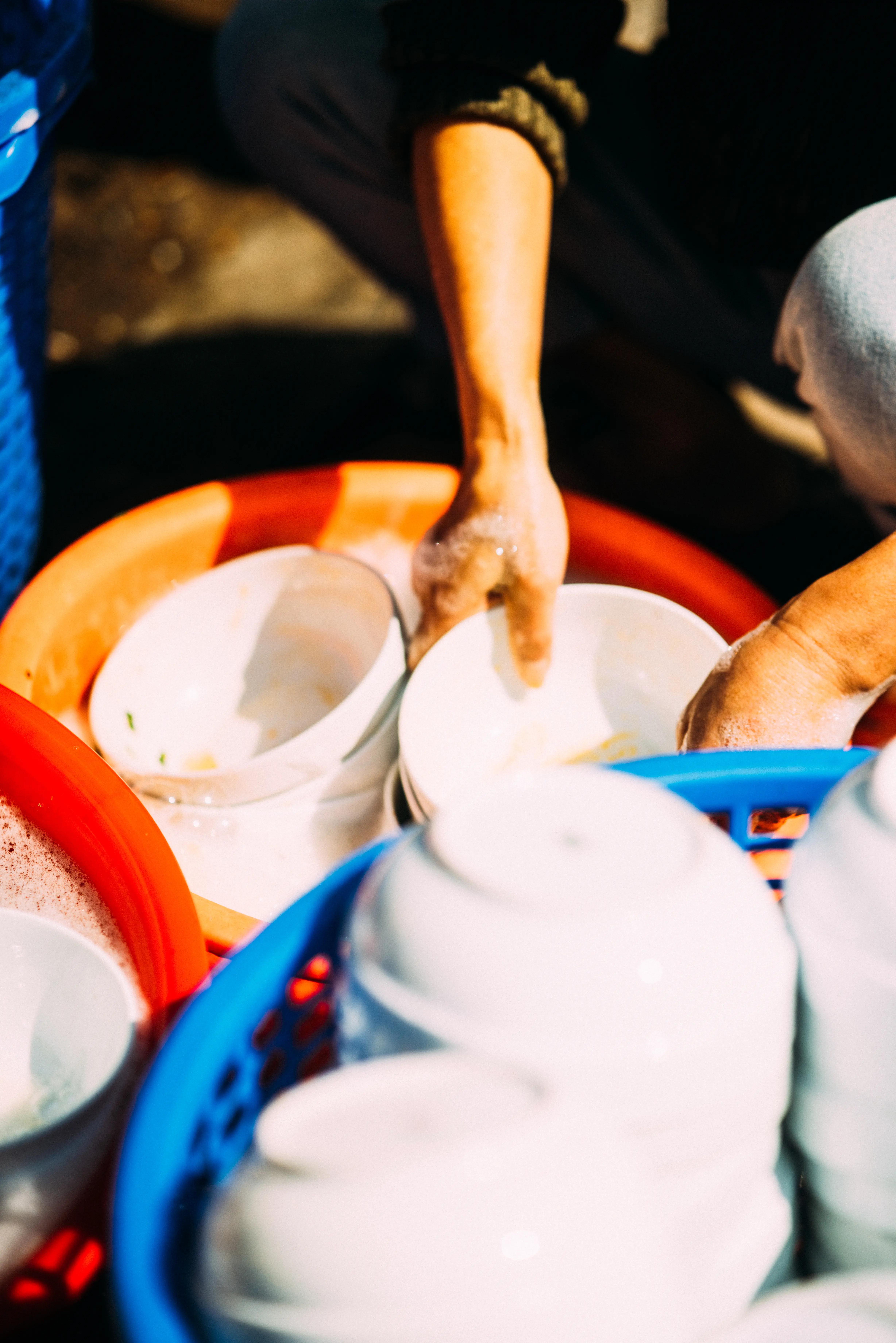 Hands washing white bowls in colourful plastic tubs on a Southeast Asian street in harsh low-angle sunlight